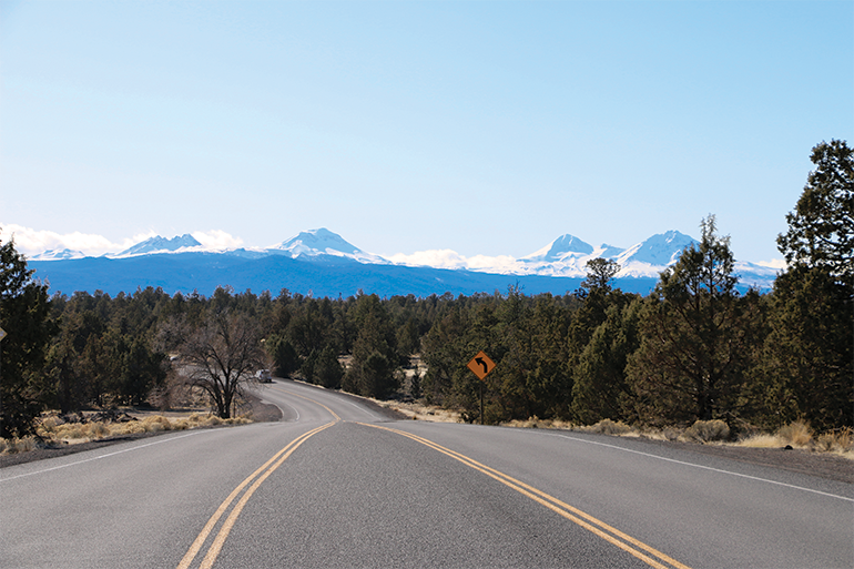 A road in Bend with coniferous trees and mountains in the background.