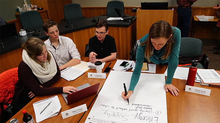 Four individuals sit around a table conversing while one takes notes on a large piece of paper.